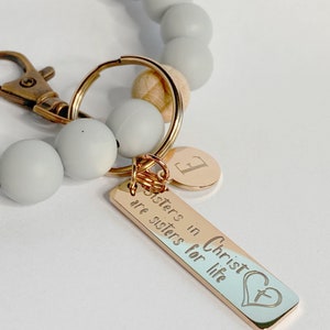 May include: A gray and wood beaded keychain with a gold charm that reads "Sisters in Christ are sisters for life" with a heart and cross design.