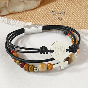 May include: Black leather bracelet with a silver clasp and a round charm with the letters "GAV" and a cross symbol. The bracelet has a strand of colorful beads and the text "Romans 8:39".