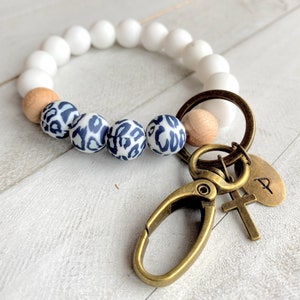 Op de afbeelding: Wit en bruin kralenarmband met een luipaardprint bedel bevestigd aan een gouden sleutelhanger met een kruis en een bedel met de letter 'P'.