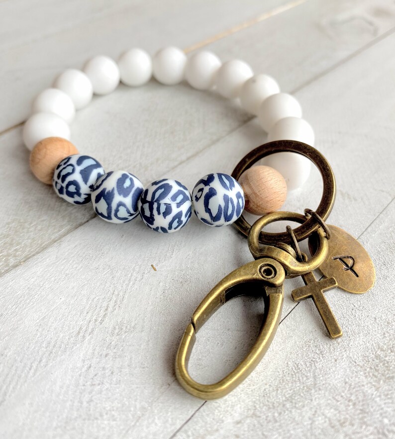 Op de afbeelding: Wit en bruin kralenarmband met een luipaardprint bedel bevestigd aan een gouden sleutelhanger met een kruis en een bedel met de letter 'P'.
