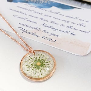 May include: A rose gold necklace with a circular pendant. The pendant contains a preserved white flower with a yellow centre, set in clear resin. The chain is fine, and a card with text is in the background.