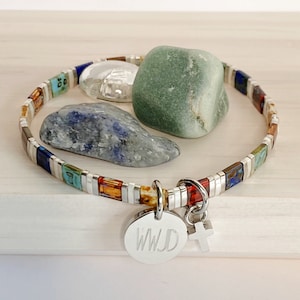 May include: A silver bracelet with colorful tiles and a charm with the letters "WJD" and a cross. The bracelet is on a wooden surface with three stones.