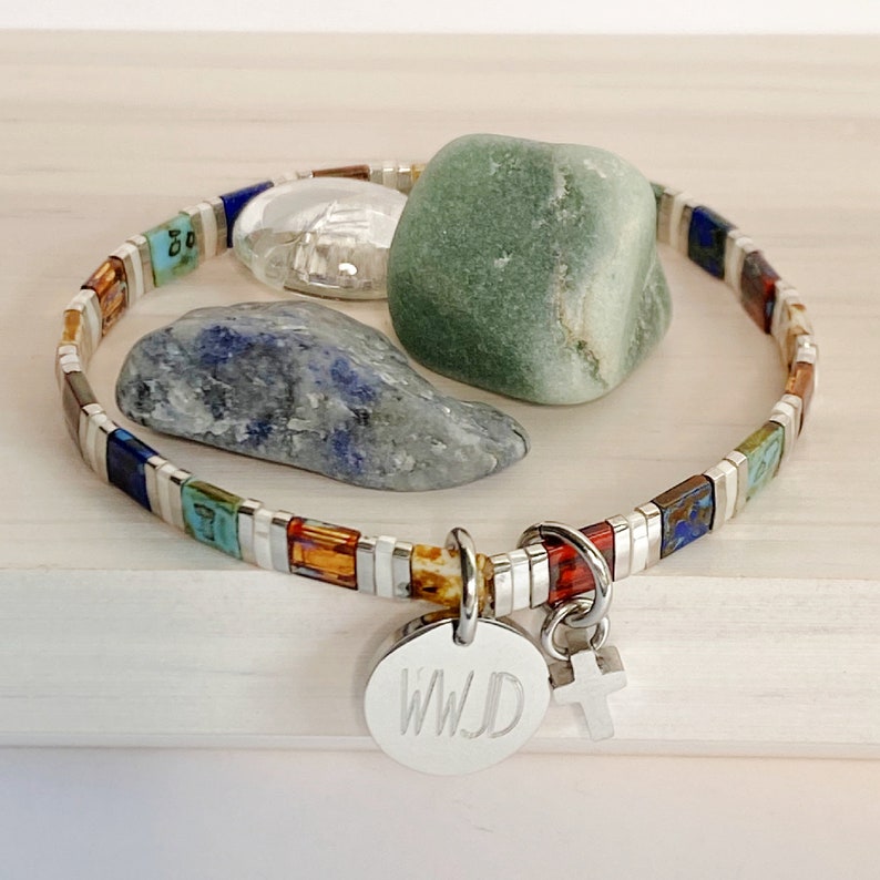 May include: A silver bracelet with colorful tiles and a charm with the letters "WJD" and a cross. The bracelet is on a wooden surface with three stones.