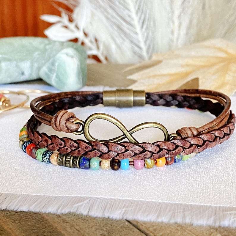 May include: A brown leather bracelet with a braided design and an infinity symbol charm. The bracelet also features colorful beads.