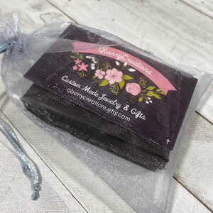 May include: A black and white card with pink floral designs and the text "Qberry Creations Custom Made Jewelry & Gifts qberrycreations.etsy.com" is inside a silver mesh bag.