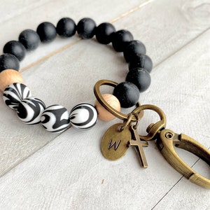Op de afbeelding: Zwart-witte kralen met een patroon, een houten kraal en een gouden bedel met de letter "W" en een kruis. De kralen zijn bevestigd aan een gouden sleutelhanger.