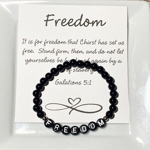 May include: A black beaded bracelet with the word "FREEDOM" spelled out in white letter beads. The bracelet is displayed on a white card with the word "Freedom" and a Bible verse printed on it.