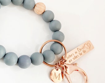May include: A gray beaded keychain with a wooden bead, a gold metal key ring, a gold metal charm with the letter M, and a gold metal tag with the text "Sisters in Christ are sisters for life" and a heart symbol.