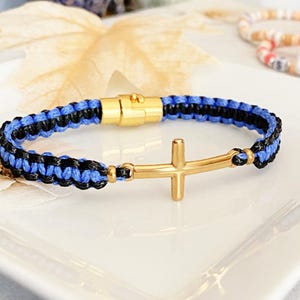 May include: A braided bracelet featuring blue and black cord with a gold-colored cross pendant. The bracelet has a gold-colored magnetic clasp. The bracelet is displayed on a white surface.