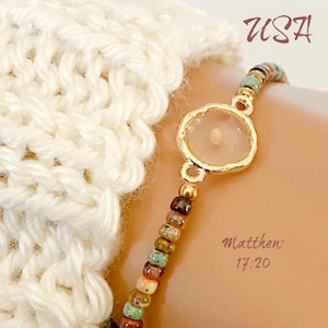 May include: A gold-toned bracelet featuring a seed encased in a clear setting. The bracelet has a beaded strand with various colors. The text "USA" and "Matthew 17:20" are visible.