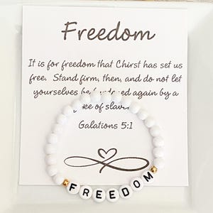 May include: White beaded bracelet with the word "FREEDOM" spelled out in white letter beads. The bracelet is displayed in front of a white card with the word "Freedom" and a Bible verse.