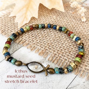 May include: A stretch bracelet featuring multicolored glass beads in shades of blue, green, red, and tan, accented with metallic bronze beads. The focal point is a bronze-toned Icthus fish charm containing a single mustard seed. Text reads: Icthus mustard seed stretch bracelet.