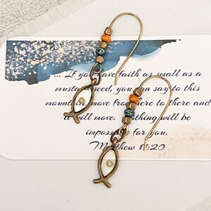 May include: A pair of dangle earrings with a fish-shaped charm. The earrings have a gold-colored hook and are adorned with colorful beads in shades of orange, blue, and brown. The earrings are displayed on a card with handwritten text.