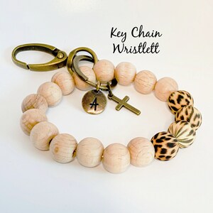 Op de afbeelding: Een houten kralen sleutelhanger armband met een gouden metalen sluiting, een bedel met de letter "A" en een kruisbedel. De armband heeft een luipaardprint op twee van de kralen.
