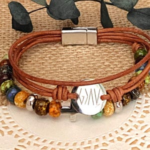 May include: A multi-strand bracelet featuring brown leather cords and a variety of colorful, textured beads in shades of green, blue, yellow, and brown. A silver-toned circular charm is engraved with the text G>AV and a silver magnetic clasp.