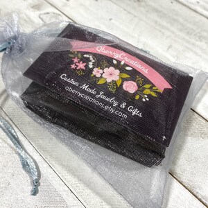 Op de afbeelding: Een kleine, zwarte cadeauzak met een bloemenontwerp en de tekst "Qberry Creations Custom Made Jewelry & Gifts qberrycreations.etsy.com" zit in een lichtblauwe organza zak.