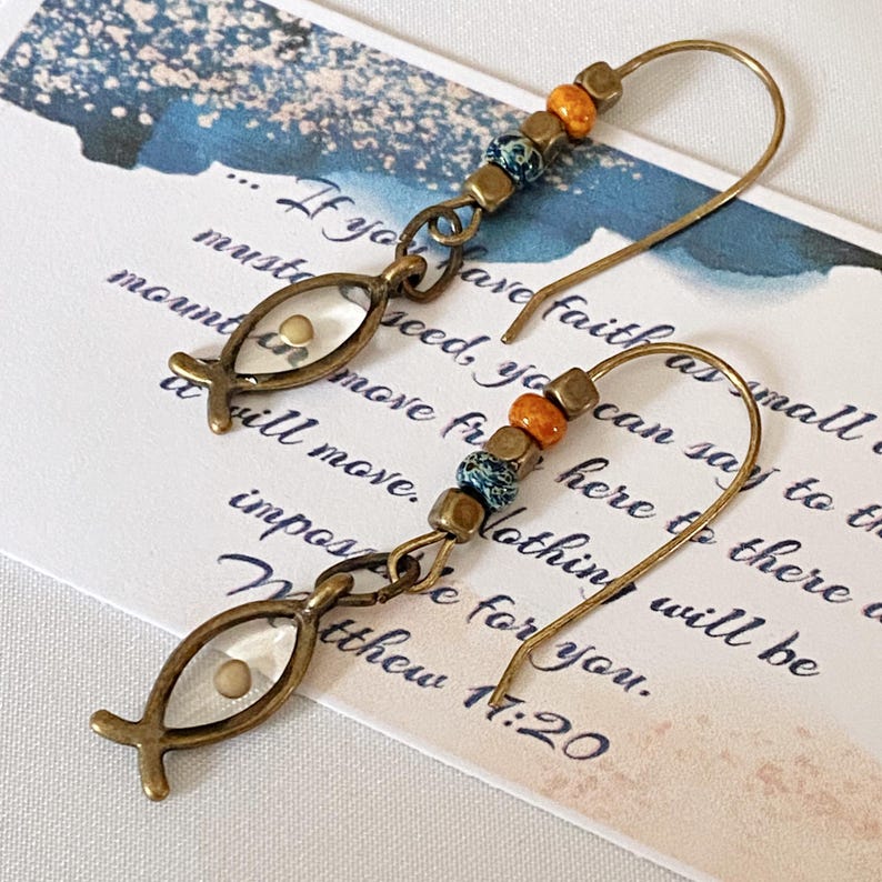 May include: A pair of handcrafted earrings featuring a fish-shaped charm with a small seed inside, connected to a beaded section with orange, blue, and gold beads. The earrings have a bronze finish and are displayed on a card with handwritten text.