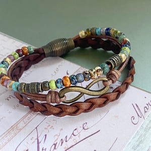 May include: A brown leather bracelet with a braided design and a multi-colored beaded strand. The bracelet features an infinity symbol charm.