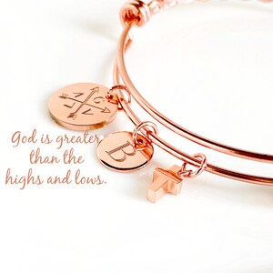 May include: Rose gold bangle bracelet with three charms: a compass, a letter 'B', and a cross. The bracelet has a quote engraved on it: "God is greater than the highs and lows."
