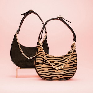 Könnte beinhalten: Zwei halbmondförmige Handtaschen. Eine ist schwarz mit einem Kettenriemen, die andere hat einen Zebra-Print mit einem goldenen Kettenriemen. Beide haben schwarze Griffe und sind vor einem rosa Hintergrund platziert.