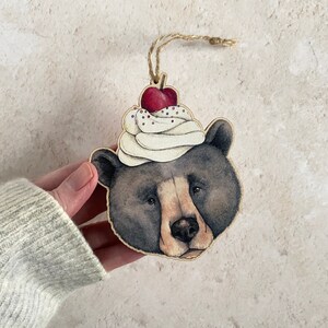 A wooden ornament of a black bear wearing a white and red cherry ice cream sundae on its head. The bear has a brown nose and black eyes. The ornament is two dimensional and is cut to the irregular shape of the animal. It has a loop of twine to hang it with.