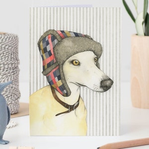 A greetings card stood on a desk next to a model penguin and a plant pot. The card features a watercolour illustration of a blonde whippet wearing a multicoloured deerstalker hat. The background behind the dog is narrow grey and white stripes.