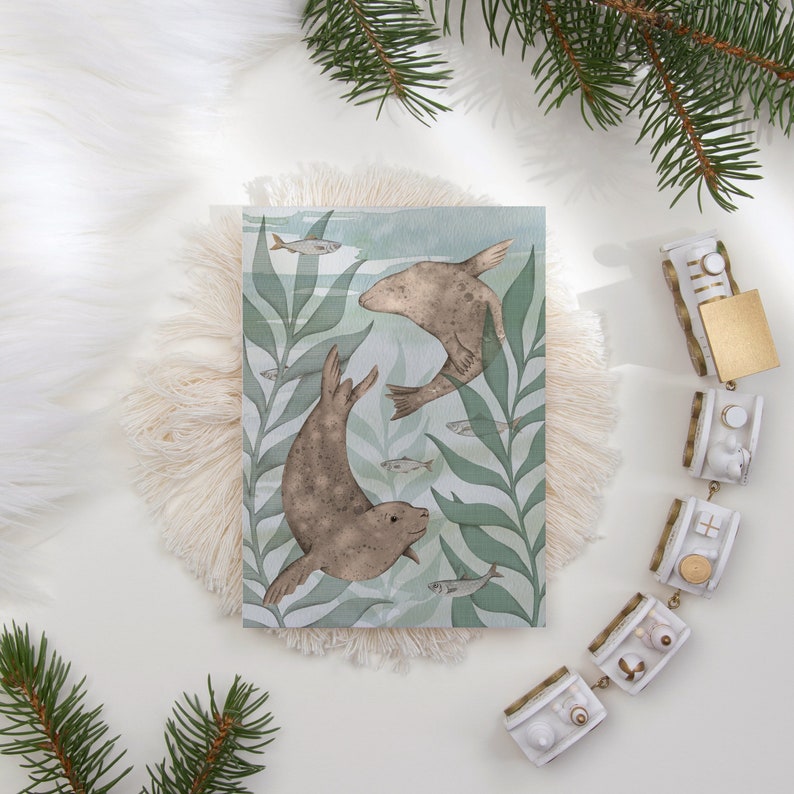 An A5 postcard with a cute illustration of two grey seals swimming through green kelp leaves with some small fish. The postcard is on a fluffy placemat next to oa toy train.