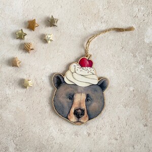 May include: A wooden ornament shaped like a bear wearing a white and red whipped cream hat with a cherry on top. The bear has brown fur and a black nose.