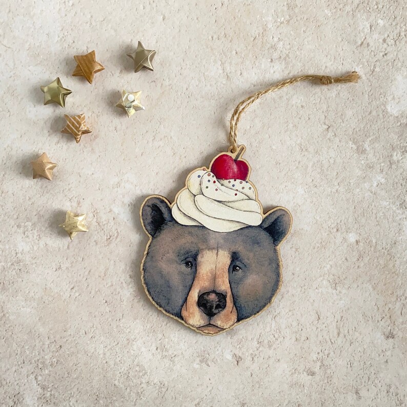 May include: A wooden ornament shaped like a bear wearing a white and red whipped cream hat with a cherry on top. The bear has brown fur and a black nose.