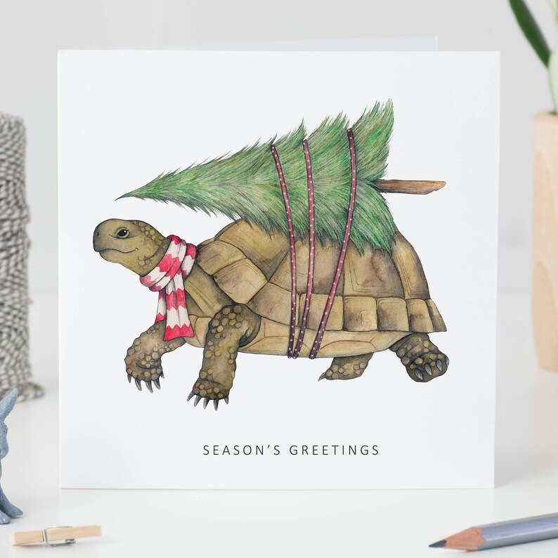 A square card stood on a desk. The card is white with a watercolour illustration of a tortoise. He is wearing a red and white striped scarf and has a Christmas tree strapped to the top of his shell. Below him, the words Season's Greetings.