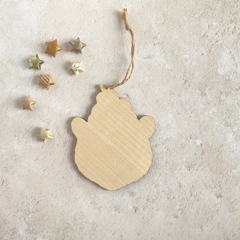 May include: A wooden ornament shaped like a bear's head with a string attached for hanging. The ornament is light brown and has a natural wood grain. It is surrounded by small gold stars.