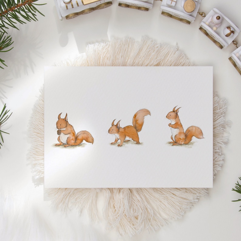 A plain white postcard with three cute red squirrels in the centre. The illustrations are detailed and have watercolour textures. The postcard is on a fluffy placemat next to a toy train.