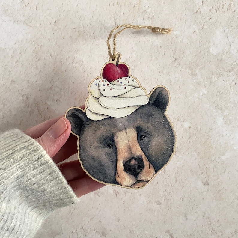A wooden ornament of a black bear wearing a white and red cherry ice cream sundae on its head. The bear has a brown nose and black eyes. The ornament is two dimensional and is cut to the irregular shape of the animal. It has a loop of twine to hang it with.