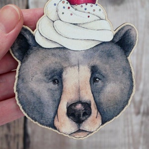 May include: A black bear with a white and red frosting cupcake on its head. The cupcake has sprinkles and a red cherry on top.