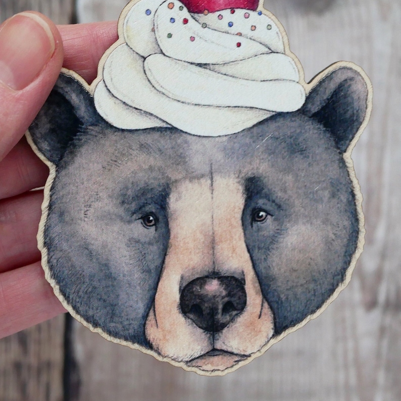 May include: A black bear with a white and red frosting cupcake on its head. The cupcake has sprinkles and a red cherry on top.