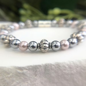 Könnte beinhalten: Ein silbernes und rosafarbenes Armband mit einem dekorativen silbernen Anhänger. Das Armband besteht aus runden Perlen und einer einzigen größeren, verzierten Perle.