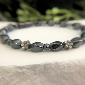 Single-strand bracelet made of 4mm black magnetic twist/swirl and round hematite beads with metallic silver-colored daisy spacer beads sitting on a white rock slab with greenery in the background. Contains a magnetic barrel clasp closure.