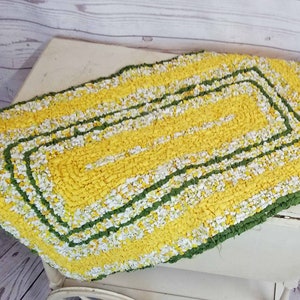 May include: A yellow and green braided rug with a white and yellow striped pattern. The rug has a rectangular shape and a fringed edge.