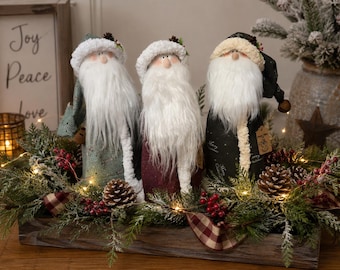 May include: Three standing Santa Claus figurines with long white faux fur beards and fabric hats. The left figure wears a sage green hat, the centre wears a white hat, and the right wears a black hat with a small brown tag attached to the side.