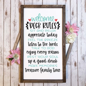 May include: A rectangular wooden sign with a dark brown frame and a white background featuring black and teal text that reads: welcome DECK RULES SIT AND RELAX appreciate today FEEL THE BREEZE listen to the birds TALK TO GOD enjoy every season REST YOUR SOUL sip a good drink MAKE MEMORIES treasure family time.