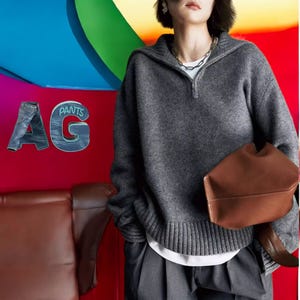 Könnte beinhalten: Ein grauer Kapuzenpullover mit Reißverschluss und Rippenbündchen. Der Pullover wird über einem weißen Hemd und einer grauen Hose getragen. Die Person hält eine braune Ledertasche.