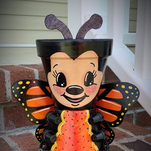 May include: A hand-painted terracotta planter shaped like a butterfly with a black head and orange wings. The planter has black dots and a yellow accent on the wings. The planter is decorated with a cute face and has a handwritten signature on the bottom.