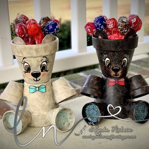 May include: Two painted terracotta flower pots shaped like bears, one brown and one white, with lollipops inside. The bears are wearing bow ties and have glitter on their feet. The text "Handmade by Lynda Nielsen" and a website address are visible below the bears.