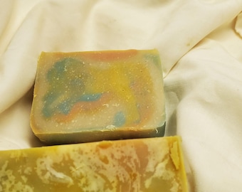 May include: Two bars of handmade soap, one with a green, blue, and yellow swirl pattern and the other with a marbled green and yellow pattern. The soap bars are sitting on a white cloth.