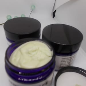 May include: Three small jars of body butter with black lids and purple glass jars. The jar in the center is open and shows the light yellow body butter inside.