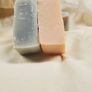 May include: Two bars of soap, one is light blue and the other is light pink. Both bars are rectangular and have a smooth surface.