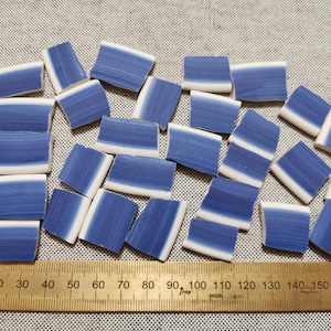 May include: A collection of small, rectangular ceramic tiles with a blue and white striped design. The tiles are arranged on a textured surface, with a wooden ruler showing measurements in centimetres.