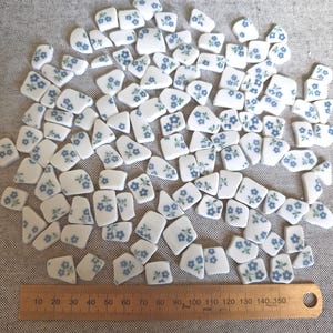 May include: A collection of white ceramic mosaic tiles featuring a blue floral pattern, arranged above a wooden ruler measuring 150 millimetres in length. Each tile piece displays a small cluster of blue flowers with green stems and leaves.