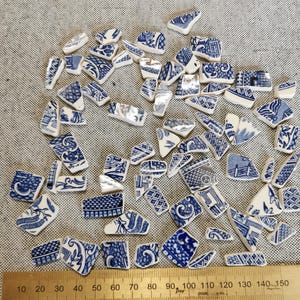 May include: A collection of blue and white ceramic shards with various patterns, including floral and geometric designs. The fragments are scattered on a textured surface, with a ruler along the bottom showing measurements in centimetres. The pieces vary in size and shape.