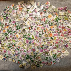 May include: A large collection of assorted ceramic shards featuring various colourful floral patterns, botanical illustrations, and green leaf motifs. These irregular, broken pieces of porcelain display a wide range of pink, yellow, blue, and green hues on white backgrounds.